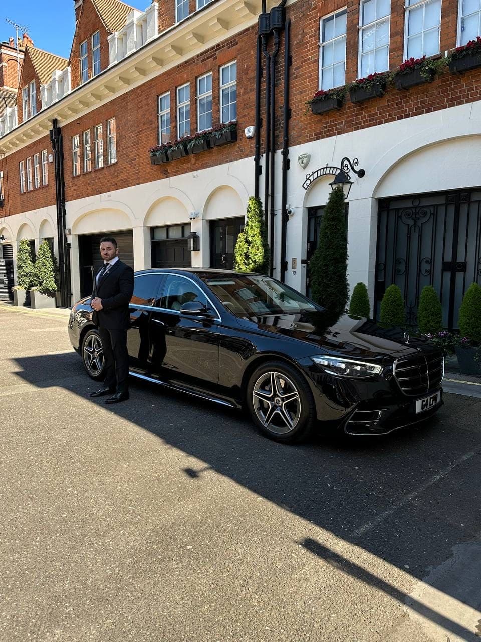 A uniformed chauffeur holding the rear door of a black Mercedes S-Class open on a London mews