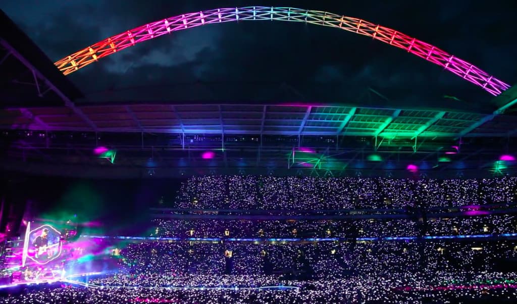 Wembley Stadium at night during a stadium concert, the arch lit in colour above a crowd wearing illuminated LED wristbands